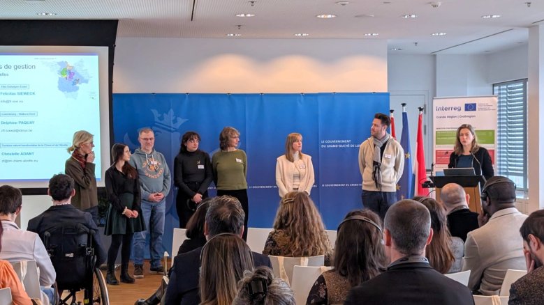 Mehrere Personen stehen nebeneinander auf einer Bühne vor einem Publikum, während eine Präsentationsfolie und ein Interreg‑Banner im Hintergrund sichtbar sind.