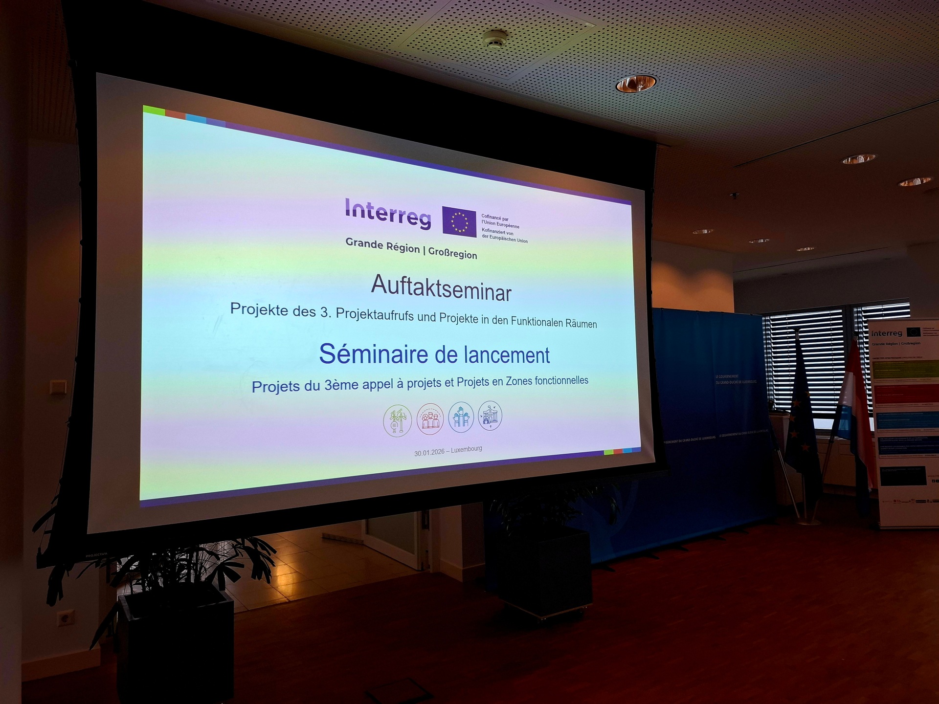 Projektionsleinwand mit der Anzeige ‚Auftaktseminar – Projekte des 3. Projektaufrufs und Projekte in den Funktionalen Räumen‘ in Deutsch und Französisch; im Raum stehen ein Interreg‑Banner und mehrere Flaggen.