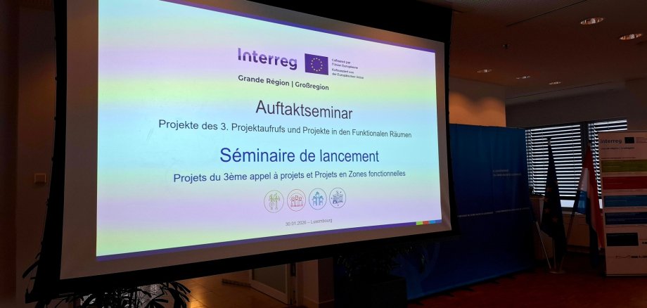 Projektionsleinwand mit der Anzeige ‚Auftaktseminar – Projekte des 3. Projektaufrufs und Projekte in den Funktionalen Räumen‘ in Deutsch und Französisch; im Raum stehen ein Interreg‑Banner und mehrere Flaggen.