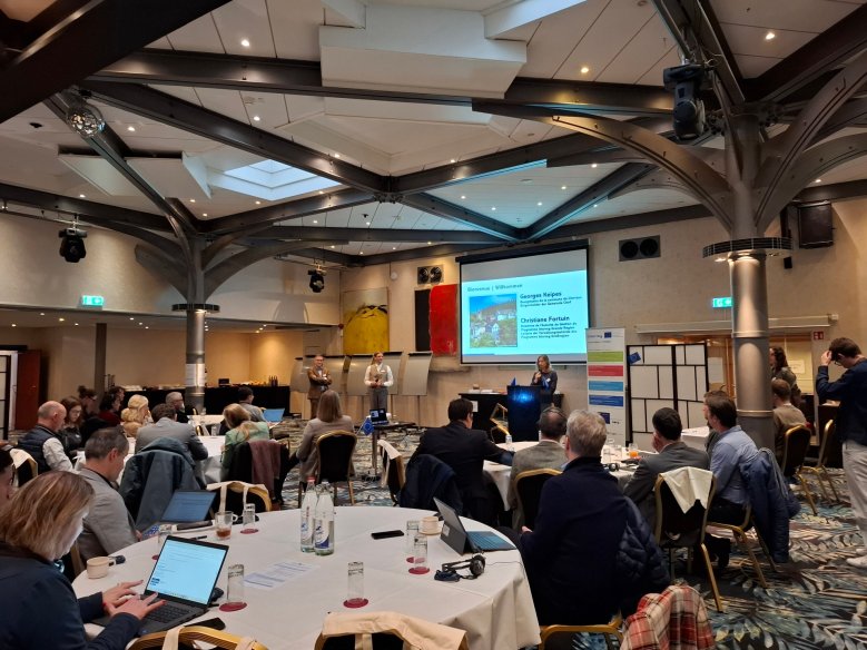 Tagungsraum mit Teilnehmenden an runden Tischen; vorne präsentiert eine Person an einem Rednerpult vor einer Leinwand