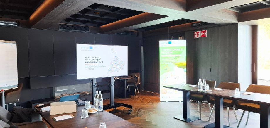 Konferenzraum mit  Tischen, auf denen Wasserflaschen, Gläser und Schreibmaterial vorbereitet sind; im Hintergrund steht ein Bildschirm für Präsentationen.