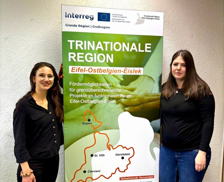 Zwei Personen stehen links(Felicitas Sieweck) und rechts(Julia Altenburg) neben einem Roll‑up‑Banner zur Trinationalen Region Eifel‑Ostbelgien‑Éislek, das Informationen zu grenzüberschreitenden Fördermöglichkeiten und eine Gebietskarte zeigt.