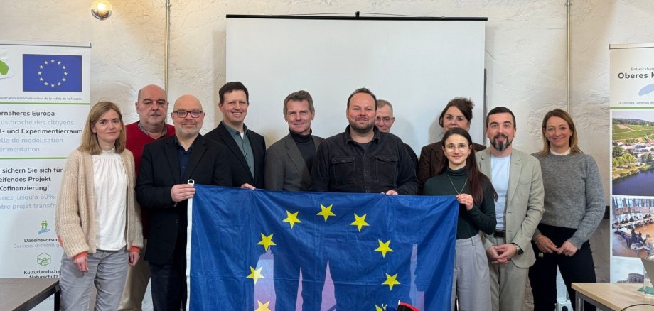 Gruppe von Personen steht in einem Seminarraum vor einer Leinwand und hält eine Europaflagge fest