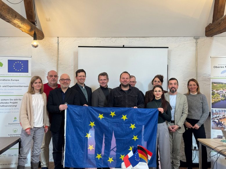 Gruppe von Personen steht in einem Seminarraum vor einer Leinwand und hält eine Europaflagge fest